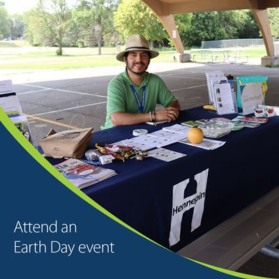Man in a hat behind a Hennepin County table with resources with text that says attend an Earth Day event