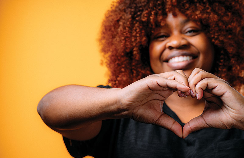 Black woman smiling at camera and making a heart shape with her hands.